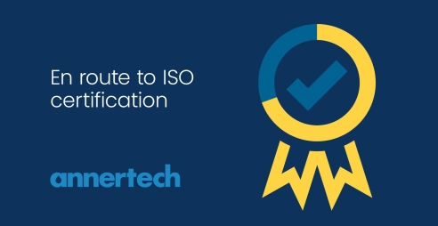 A blue and yellow graphic of a ribbon with a tick in the middle. Text next to it reads “en route to ISO certification“.