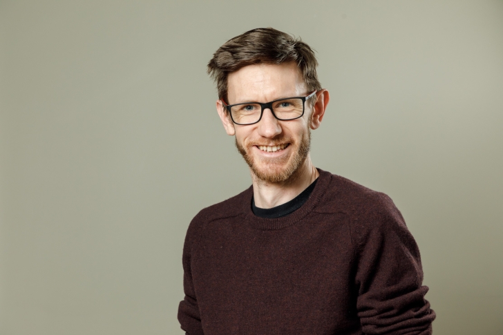 A portrait of Tom Bamford.  He's wearing glasses and a brown jersey.