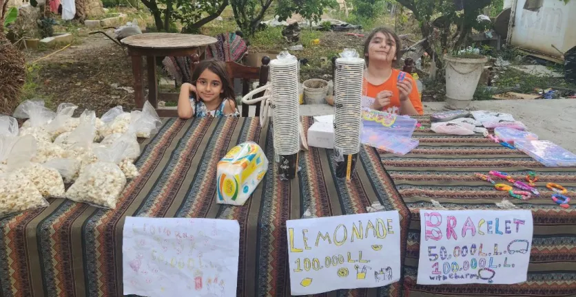 Two girls sit at a market stall selling popcorn and lemonade.