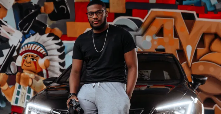 Sav Monzac sits on the bonnet of a car. Behind him is a colourful graffitied wall.