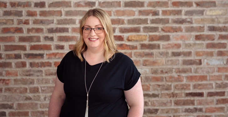 Stella Power is wearing glasses and a black top. She is standing in front of a brick wall.