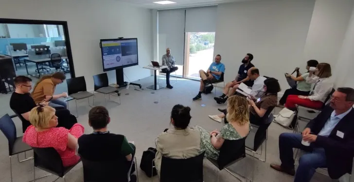 People sit on chairs in a circle, watching a presentation on a big screen.