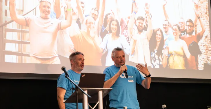 Tony Barker and Anthony Lindsay address the LocalGov Drupal Camp attendees. Behind them is a huge photo of the Annertech team.