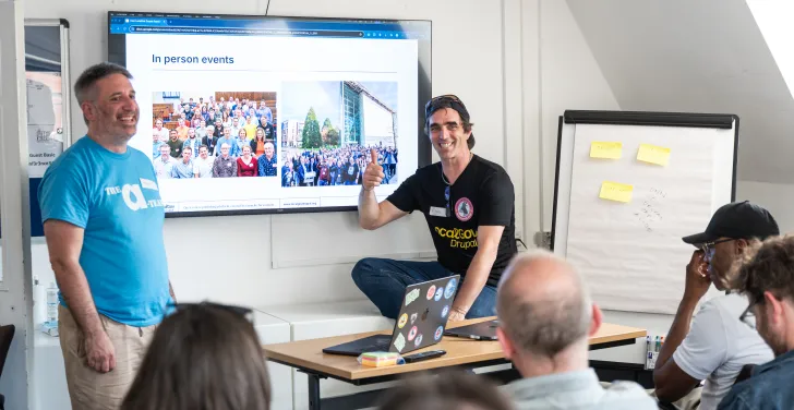 Tony Barker stands next to a table, and Finn Lewis is sitting on the table, presenting a session for LocalGov Drupal Camp 2025.