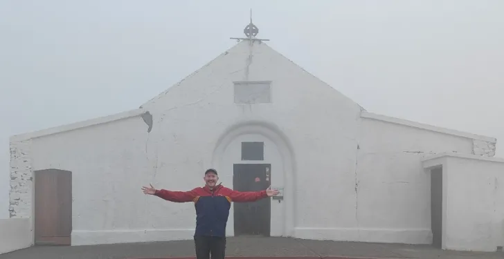 A man stands in the mist with his arms open. Behind him is a church.