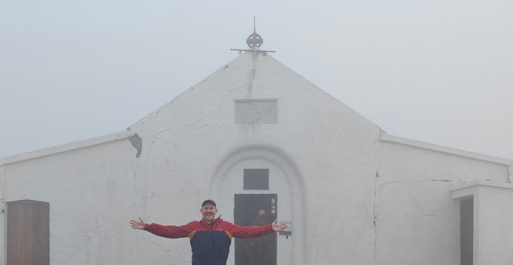 A man stands in the mist with his arms open. Behind him is a church.