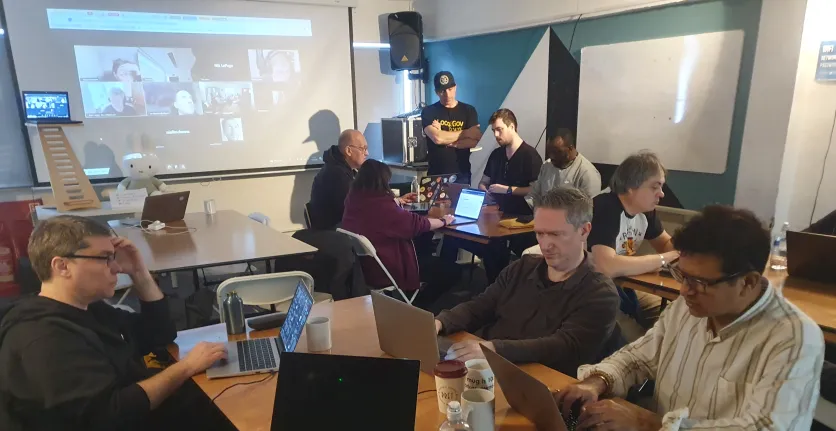 People work on their computers while other connect to the room remotely on a big screen in the background.