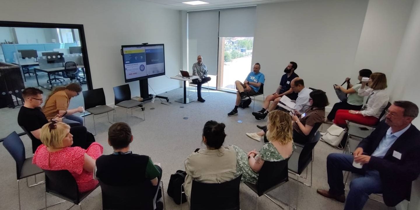 People sit on chairs in a circle, watching a presentation on a big screen.