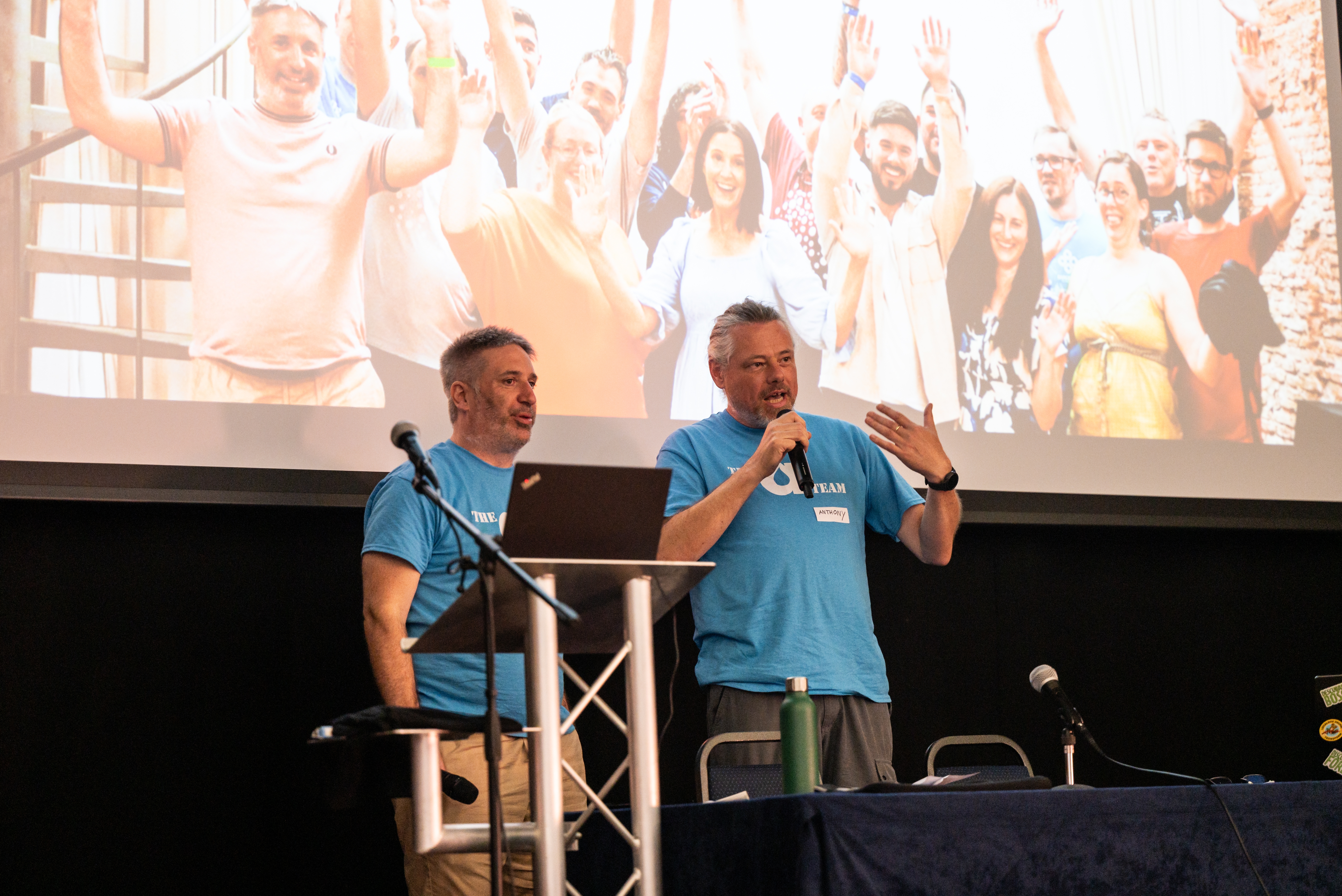 Tony Barker and Anthony Lindsay address the LocalGov Drupal Camp attendees. Behind them is a huge photo of the Annertech team.