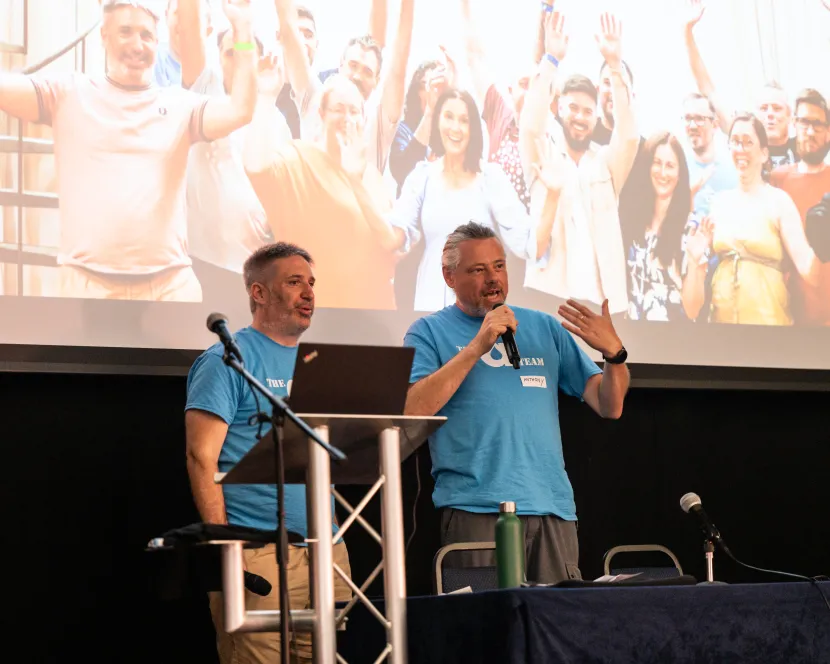 Tony Barker and Anthony Lindsay address the LocalGov Drupal Camp attendees. Behind them is a huge photo of the Annertech team.